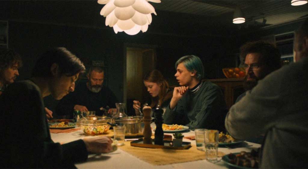 Os personagens estão sentados à mesa jantando e conversando.