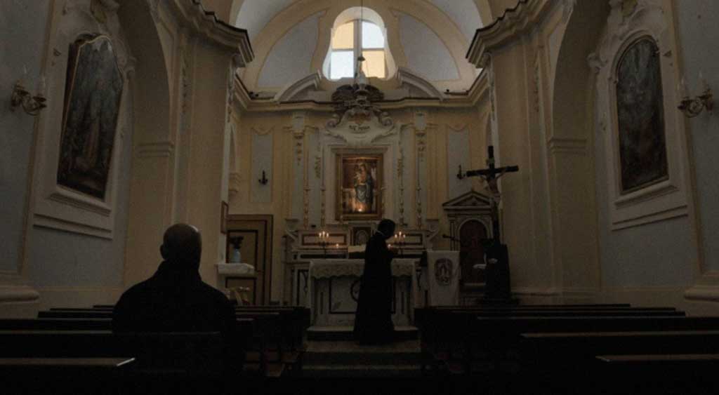 McCall sentado no banco da igreja, em frente ao altar, onde encontra-se um padre.