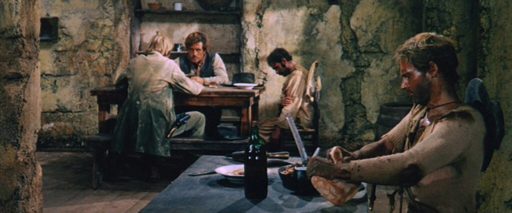 Trinity comendo pão com feijão enquanto é observado por dois pistoleiros na mesa ao fundo.