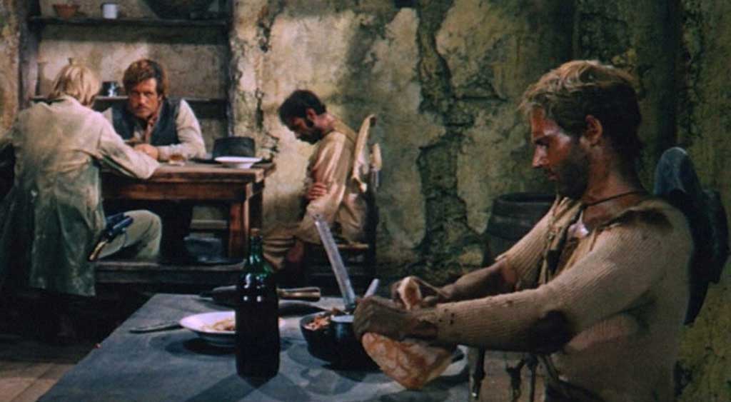 Trinity comendo pão com feijão enquanto é observado por dois pistoleiros na mesa ao fundo.