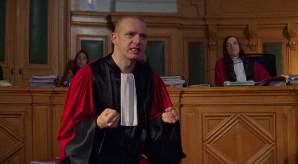 O promotor gesticula veementemente em sua fala, sendo observado ao fundo pela juíza do caso.