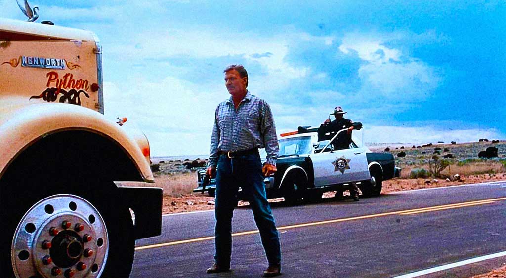 Claude Sams em pé com a arma em punho encara de frente o caminhão amarelo onde está Python, ao fundo ele é observado por um policial.