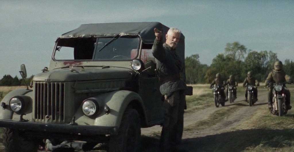 Yeagor Dragunov dá sinal com as mãos para os motoqueiros russos continuarem em frente para alcançar Korpi dirigindo seu caminhão.